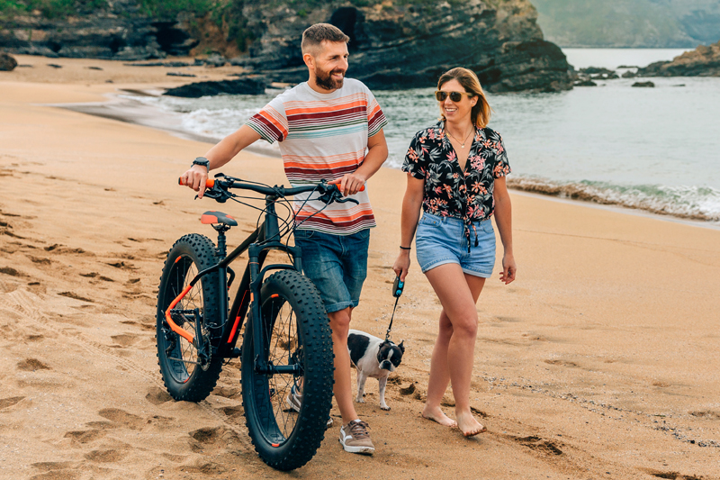 Una pareja camina por la playa con su perro y una fat-bike